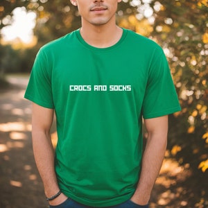 Könnte beinhalten: Ein grünes T-Shirt mit dem Aufdruck "CROCS AND SOCKS" in weiß auf der Vorderseite.