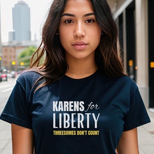 May include: A navy blue t-shirt featuring the text "Karens for Liberty" and "Threesomes Don't Count" in white and gold. The shirt is worn by a person outdoors.