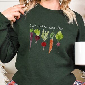 Op de afbeelding: Een donkergroen sweatshirt met de tekst "Let's root for each other" en een aquarel-illustratie van vijf groenten: een paarse wortel, een rode biet, een groene biet, een oranje wortel en een rode biet.