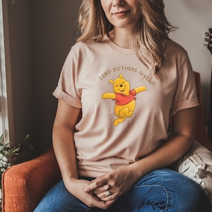 May include: A light pink t-shirt with a cartoon image of Winnie the Pooh with the text "Zero Bothers Given" in gold lettering.