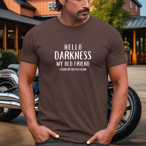 May include: A brown t-shirt with white text that reads "Hello Darkness My Old Friend I Stood Up Too Fast Again".