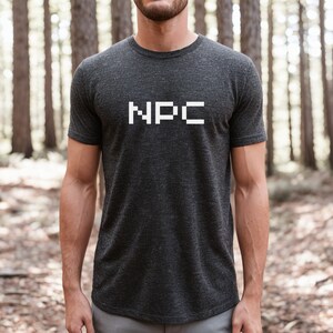 May include: Dark grey t-shirt featuring the white pixelated letters "NPC". The short-sleeved top is made of a soft material. The background is a blurred forest scene. Suitable for casual wear.