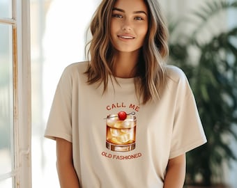 Peut inclure: Un t-shirt beige avec le texte "Call me old fashioned" et un dessin d'un cocktail old fashioned avec une cerise.