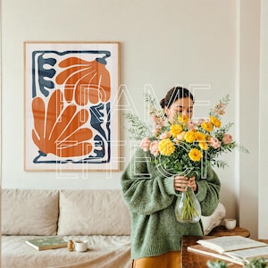 Frame Mockup with Woman in Artistic Minimalist Home – A serene, artistic interior features a woman placing pastel flowers in a vase while a large poster mockup with green edges awaits artwork on the wall behind her. The setting is complete with a soft sofa, book, and coffee cup, highlighted by natural daylight.