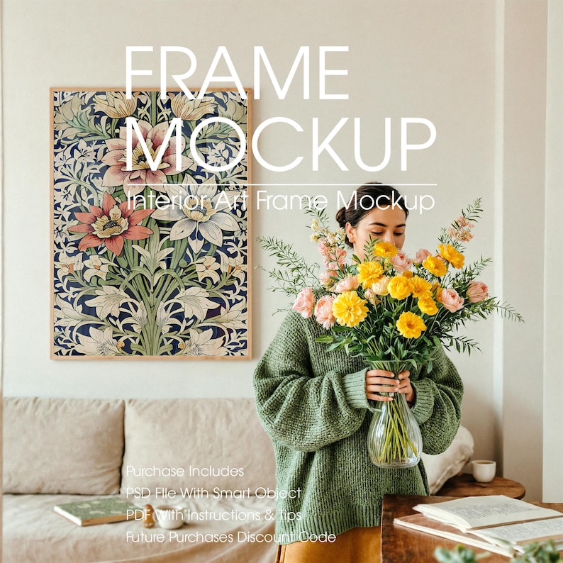 Woman with Poster Frame Mockup in Serene Minimalist Living Room – A young woman with dark hair tied back, wearing an oversized muted green sweater, arranges pastel pink and yellow flowers in a glass vase. Behind her, a large empty wooden poster frame mockup with a soft green border hangs on the wall, ready for artwork. The scene includes a linen-covered sofa, a coffee cup, and an open book on a wooden table, all bathed in soft natural light.