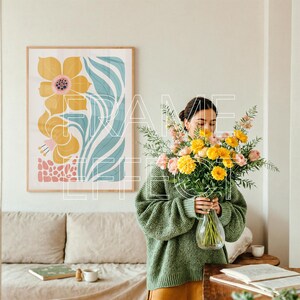 Hands Holding Mockup Scene with Woman Styling Flowers Nearby – A minimalist interior features a woman in a relaxed pose arranging pastel florals near a framed art mockup hanging above a neutral-toned sofa. The wooden frame mockup with green edging adds subtle color, while the relaxed ambiance is enhanced by a coffee cup and soft natural light.