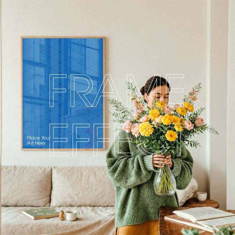 Framed Poster Mockup with Woman Styling Flowers in Cozy Interior – In a warm, neutral-toned room, a woman in a muted green sweater gently arranges flowers in a vase. A large artwork frame mockup with a subtle green border hangs above a cozy sofa. The scene includes a book and coffee on a table, emphasizing a calm and creative atmosphere.