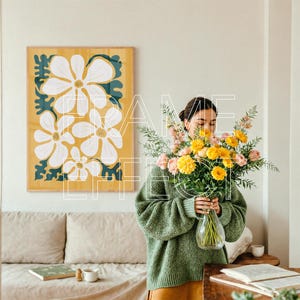 Poster Frame Mockup with Woman and Warm Interior Styling – A young woman stands in a softly lit, modern room with a bouquet of pink and yellow flowers. A wooden poster frame mockup with green trim hangs above a neutral couch, creating an inviting setting that blends relaxed styling with creative energy.