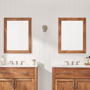 May include: Two matching bathroom vanities with wooden cabinets, white countertops, and brushed nickel faucets. Each vanity has a rectangular mirror with a wooden frame above it and a glass wall sconce with a clear glass shade.
