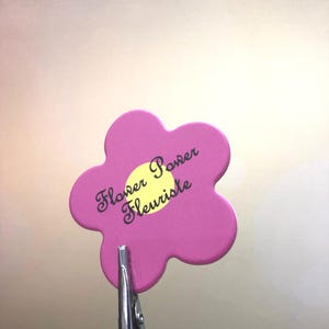 May include: Pink flower-shaped clip with the text "Flower Power Fleuriste" in black lettering. The flower has a yellow center.