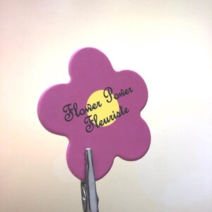 May include: A pink flower-shaped sign with the text "Flower Power Fleuriste" in black cursive writing. The sign is attached to a silver clip.