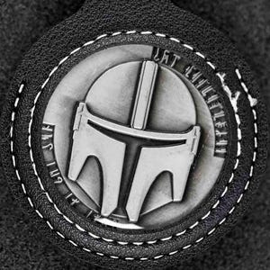 May include: A black leather keychain with a silver Mandalorian helmet emblem. The emblem is circular with the words "This is the way" around the edge. White stitching outlines the emblem and the keychain.