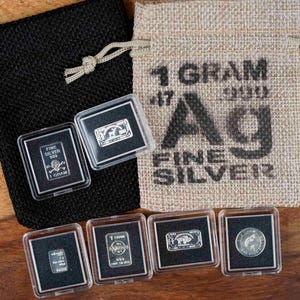 Puede incluir: Una variedad de lingotes y monedas de plata fina en estuches protectores transparentes, junto con una bolsa negra con cordón y una bolsa de yute. La bolsa de yute está impresa con "1 GRAM .999 FINE SILVER". Las piezas de plata presentan varios diseños.