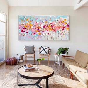 May include: A large abstract painting with a white background and colorful flowers in shades of pink, orange, yellow, and blue. The painting is hanging on a white wall in a living room setting with furniture and plants.
