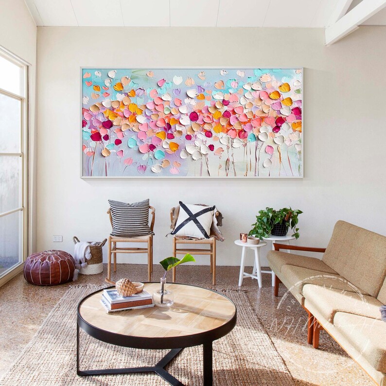 May include: A large abstract painting with a white background and colorful flowers in shades of pink, orange, yellow, and blue. The painting is hanging on a white wall in a living room setting with furniture and plants.