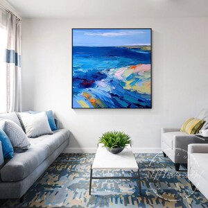May include: Abstract painting of a seascape with blue, green, and white colors. The painting is framed in black and hangs on a white wall above a gray couch.