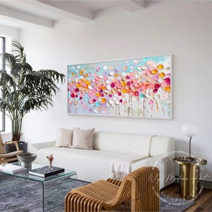 May include: Abstract painting with a colorful floral design. The painting features a variety of pink, orange, yellow, and blue flowers on a light blue background. The painting is framed in a white frame and is hanging on a white wall.