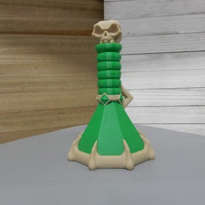 May include: A green and beige bottle stopper with a skull on top. The bottle stopper has a diamond-shaped ring around the neck and a base with claw-like feet.