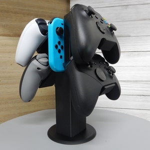 May include: A black stand with four controllers: a white Nintendo Switch Joy-Con, a gray Nintendo Switch Joy-Con, and two black Xbox controllers.