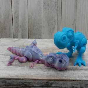 Puede incluir: Dos pequeños juguetes de dinosaurio, uno azul y otro morado, están colocados sobre una superficie de madera blanca. El dinosaurio azul está de pie, mientras que el dinosaurio morado está tumbado de lado.