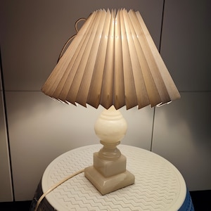 May include: A white alabaster table lamp with a pleated fabric shade. The shade is made of multiple layers of fabric that are folded and sewn together to create a pleated effect. The lamp is lit and casting a warm glow.