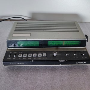 Può includere: Un radio-sveglia elettronico vintage argento e nero con un display digitale verde e una varietà di pulsanti etichettati in tedesco. Il radio-sveglia è etichettato "electronic radio timer" e "UNIVERSUM".