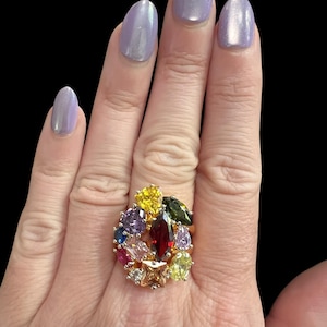 May include: A gold-toned ring featuring a cluster of multi-colored faceted gemstones in various shapes, including marquise, pear, and round cuts. The stones are arranged in a dense, floral-inspired pattern with red, yellow, purple, blue, and green hues.