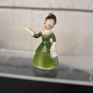 May include: A vintage ceramic figurine of a woman in a green dress with a matching hat and a white purse. The figurine has a detailed face with closed eyes and red lips. The dress has a flared skirt and a white collar.