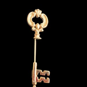 Op de afbeelding: Een goudkleurige sleutelspeld met een sierlijk ontwerp. De speld heeft een sleutelvorm met een decoratieve bovenkant en een lange, slanke speld. De details van de sleutel omvatten een afgeronde bovenkant, een centraal ontwerp en een sleutelgatvorm.