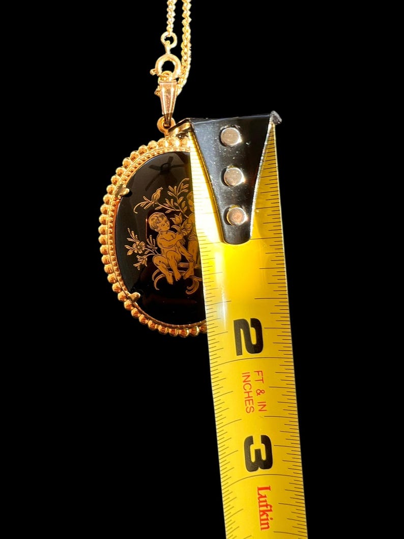 Peut inclure: Collier pendentif dor&eacute; avec un centre ovale noir orn&eacute; d'un motif de ch&eacute;rubin dor&eacute;. Le pendentif mesure environ 5 cm de long, avec une bordure perl&eacute;e et une cha&icirc;ne d&eacute;licate.
