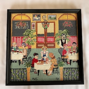 May include: Framed needlepoint artwork depicting a cafe scene with patrons and staff. The scene features tables, chairs, and a bar area, with the text "CAFE" visible. The artwork is framed in a black frame and has a detailed, colorful design.