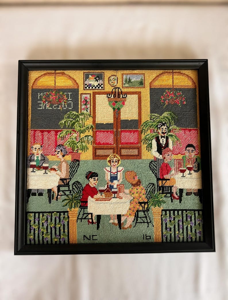 May include: Framed needlepoint artwork depicting a cafe scene with patrons and staff. The scene features tables, chairs, and a bar area, with the text "CAFE" visible. The artwork is framed in a black frame and has a detailed, colorful design.
