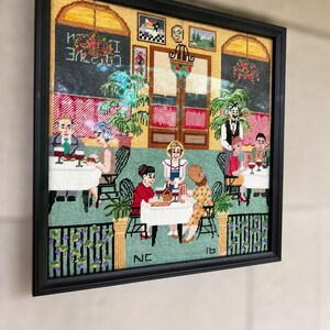 May include: Framed needlepoint art depicting a restaurant scene with patrons and staff. The artwork features a black frame, a green and red color scheme, and the text "Cuisine Italienne" in the upper left corner. The scene includes tables, chairs, and decorative elements.