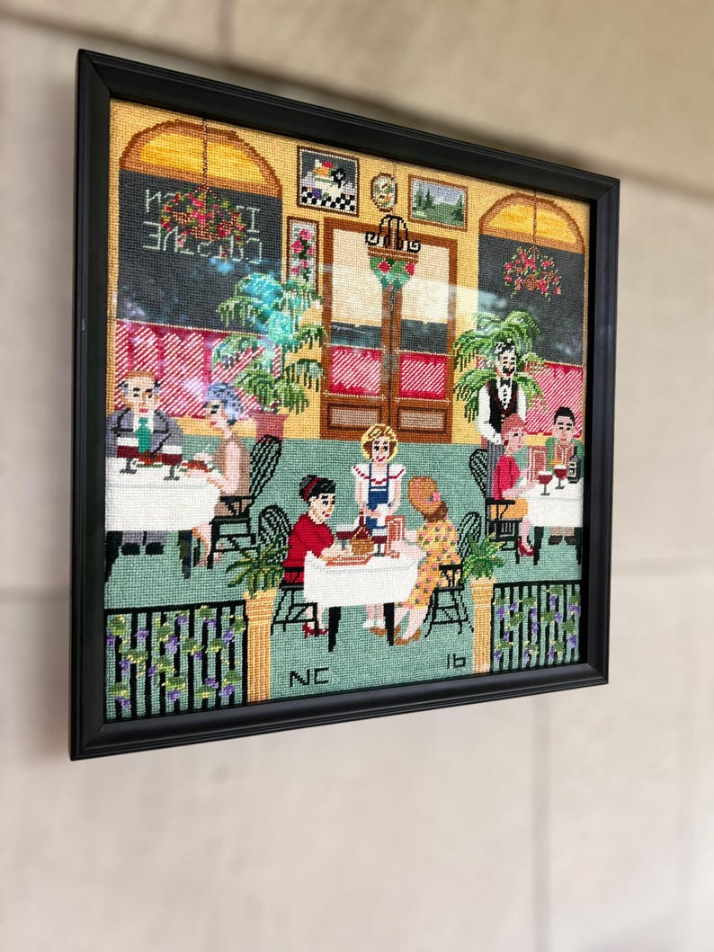 May include: Framed needlepoint art depicting a restaurant scene with patrons and staff. The artwork features a black frame, a green and red color scheme, and the text "Cuisine Italienne" in the upper left corner. The scene includes tables, chairs, and decorative elements.