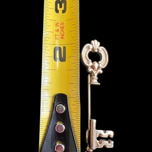 Op de afbeelding: Een goudkleurige sleutelspeld, ongeveer 6,35 cm lang, met een decoratieve fleur-de-lis top en een gedetailleerde sleutelbaard. De sleutel staat tegen een zwarte achtergrond, met een gele meetlint voor de schaal.