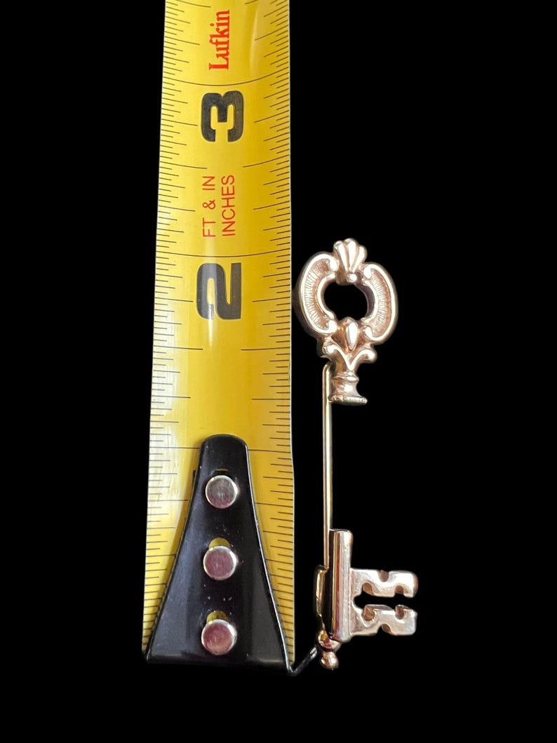Op de afbeelding: Een goudkleurige sleutelspeld, ongeveer 6,35 cm lang, met een decoratieve fleur-de-lis top en een gedetailleerde sleutelbaard. De sleutel staat tegen een zwarte achtergrond, met een gele meetlint voor de schaal.