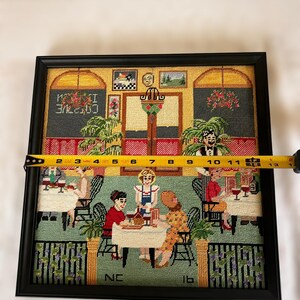 May include: Framed needlepoint artwork depicting a cafe scene with patrons and servers. The artwork is in a black frame and measures approximately 12 inches wide. The scene includes tables, chairs, and decorative elements, with the word "COTTVE" visible.