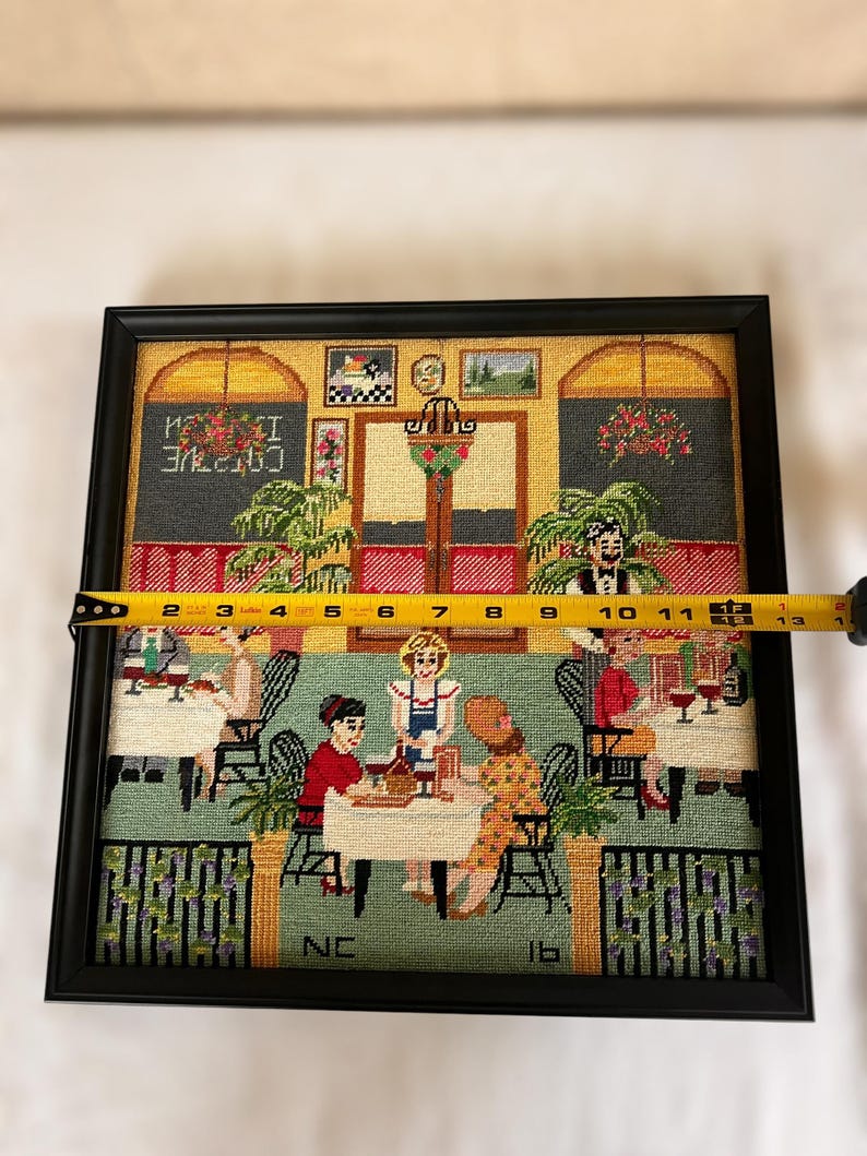 May include: Framed needlepoint artwork depicting a cafe scene with patrons and servers. The artwork is in a black frame and measures approximately 12 inches wide. The scene includes tables, chairs, and decorative elements, with the word "COTTVE" visible.