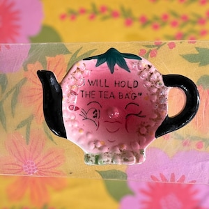 May include: A pink ceramic tea bag holder shaped like a teapot, with black handle and spout. The holder has a winking face and the text "I WILL HOLD THE TEA BAG". The background is a floral pattern.