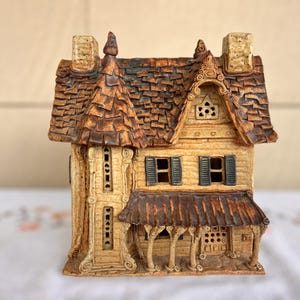 May include: A small, hand-crafted ceramic house with a brown roof, white walls, and brown accents. The house has a porch with a brown awning and a small window.