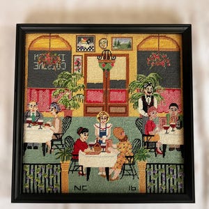 May include: Framed needlepoint artwork depicting a restaurant scene. The scene includes patrons at tables, a waiter, and the text "CAFE" in reverse. The artwork is framed in a black frame.