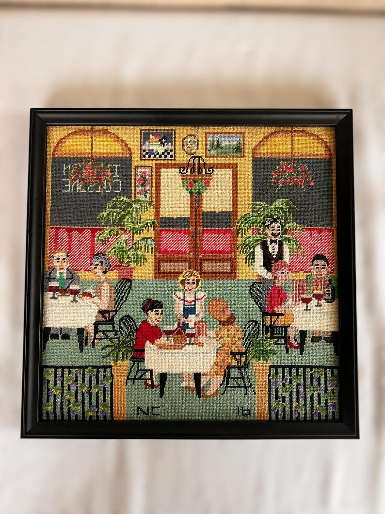 May include: Framed needlepoint artwork depicting a restaurant scene. The scene includes patrons at tables, a waiter, and the text "CAFE" in reverse. The artwork is framed in a black frame.