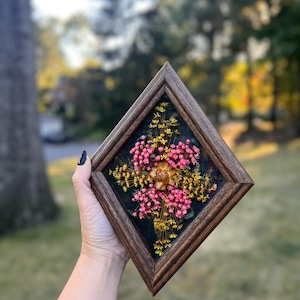 May include: A framed floral arrangement featuring pink and yellow dried flowers. The square wooden frame has a dark brown finish. The flowers are arranged in a symmetrical pattern against a dark background.