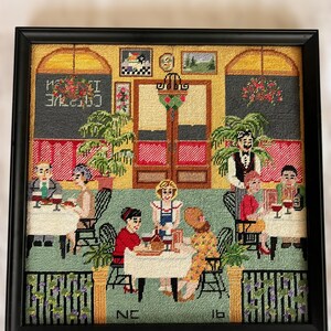 May include: Framed needlepoint art depicting a cafe scene with patrons at tables. The artwork features a black frame, a green floor, and a yellow wall. The text "CAFE" is visible. The scene includes tables, chairs, and a waiter.