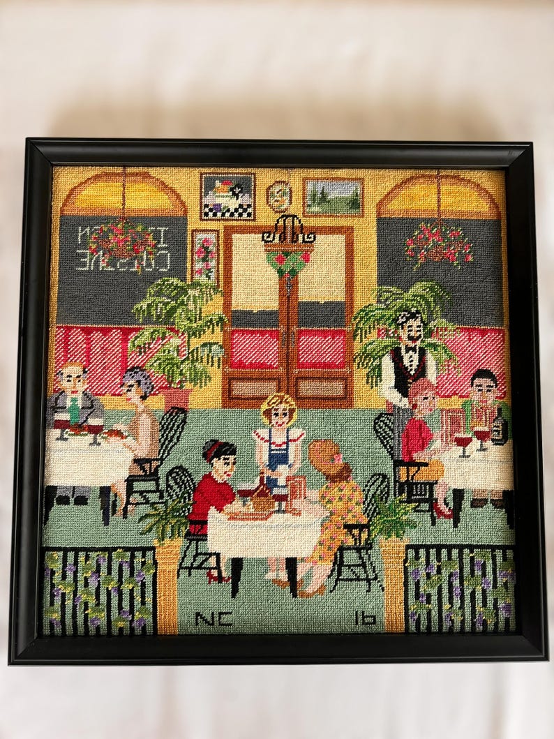 May include: Framed needlepoint art depicting a cafe scene with patrons at tables. The artwork features a black frame, a green floor, and a yellow wall. The text "CAFE" is visible. The scene includes tables, chairs, and a waiter.