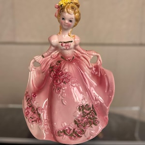 May include: A vintage porcelain figurine of a woman in a pink gown. The dress is adorned with rose accents. The woman has blonde hair and is holding the edges of her dress. The figurine is a decorative item.