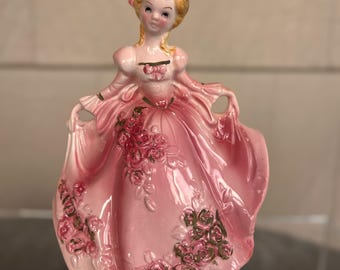 May include: A vintage porcelain figurine of a woman in a pink gown. The dress is adorned with rose accents. The woman has blonde hair and is holding the edges of her dress. The figurine is a decorative item.