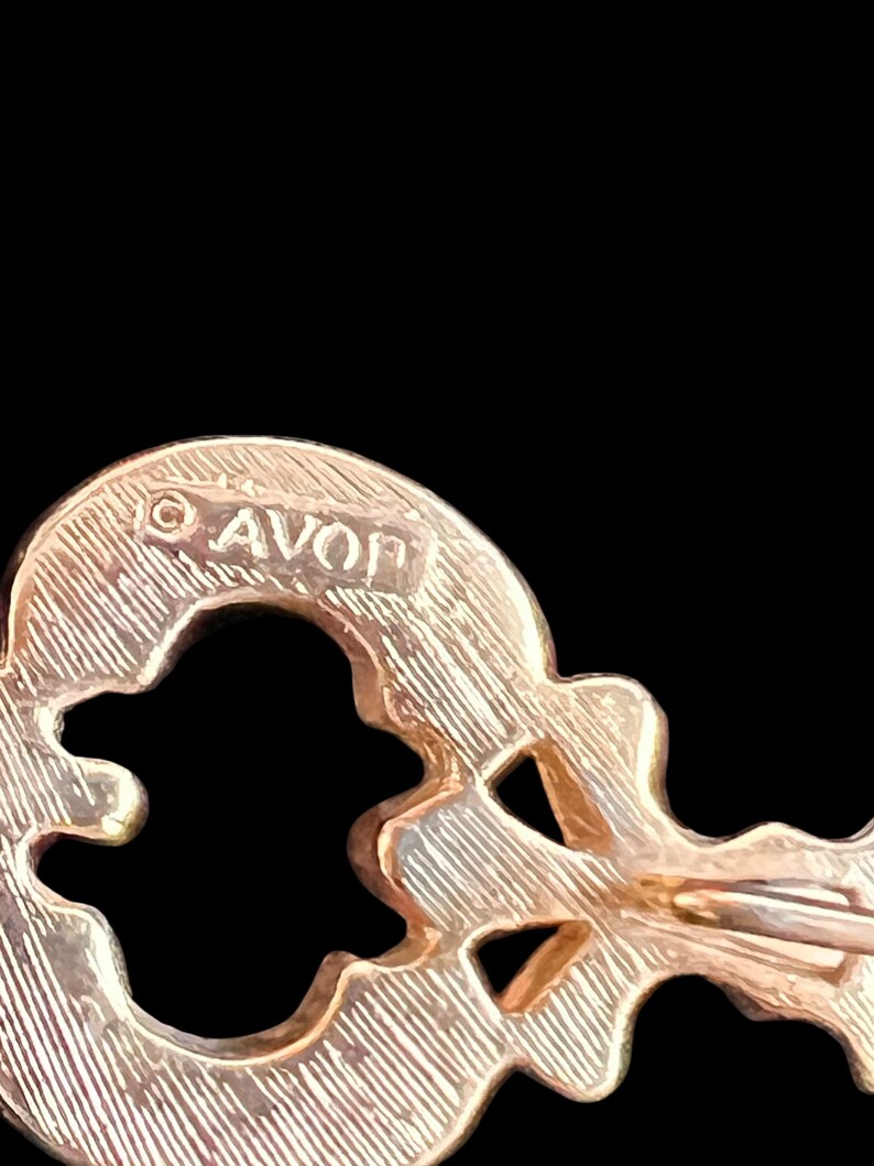 Op de afbeelding: Close-up van een goudkleurige sleutelvormige broche met het woord "AVON" op de bovenkant gestempeld. De broche heeft een getextureerd oppervlak en een sluiting met speld aan de achterkant. De achtergrond is zwart.