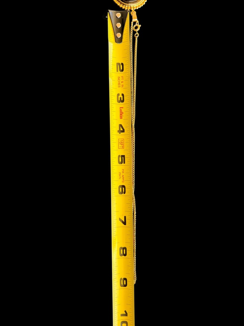 Peut inclure: Un collier dor&eacute; avec un pendentif, mesur&eacute; contre un m&egrave;tre ruban jaune. La cha&icirc;ne s'&eacute;tend sur environ 20 cm. Le pendentif a un centre sombre avec une bordure dor&eacute;e d&eacute;corative. Le fond est noir.