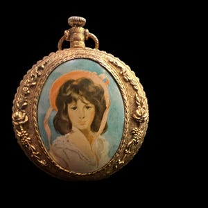 May include: A gold-toned metal pocket watch case featuring an oval portrait of a person wearing a peach-colored bonnet. The frame is decorated with embossed floral patterns and a winding crown at the top, set against a solid black background.